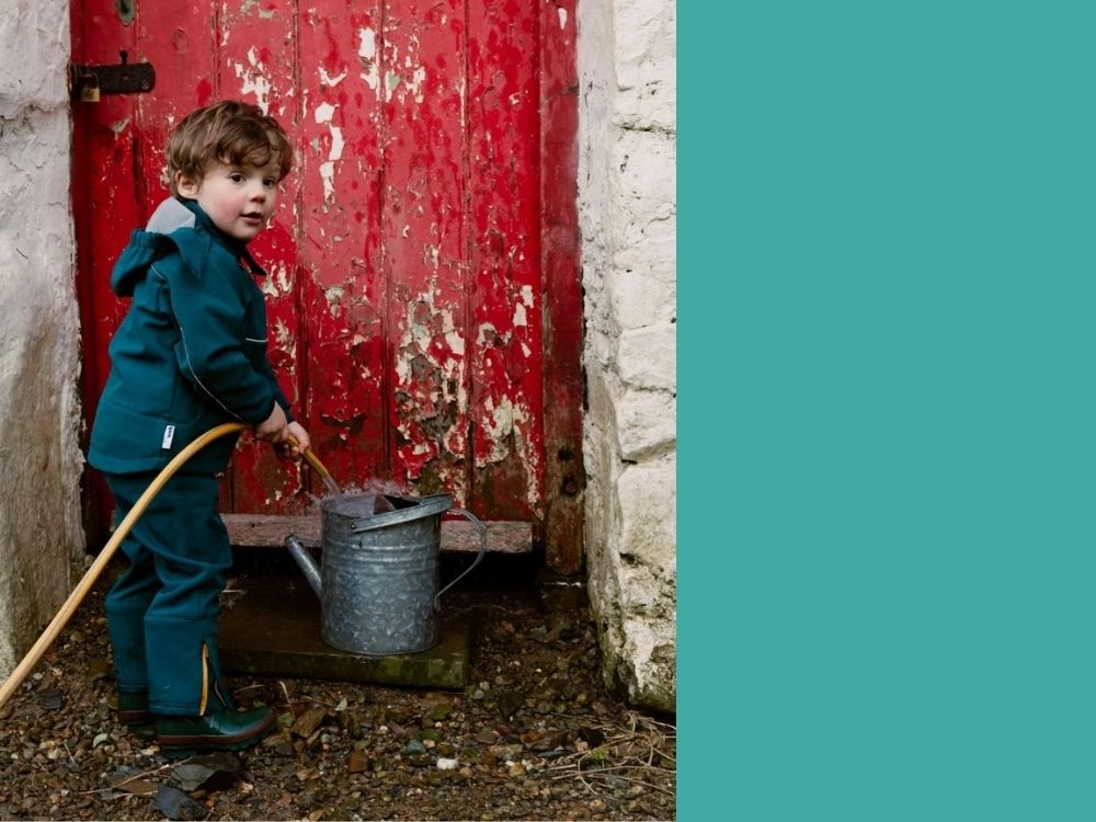 Little boy in Kidunk Clothing playing with watering can outside a red door